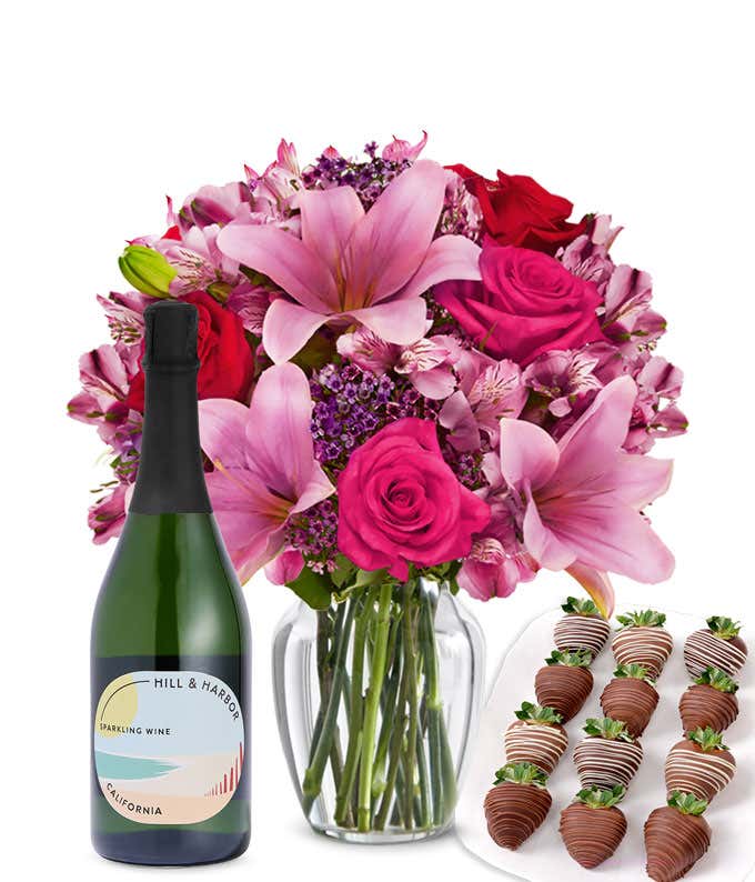 A mixed bouquet of pink and purple flowers in a clear glass vase is displayed with a bottle Hill & Harbor Sparkling Wine and 12 chocolate-covered strawberries.