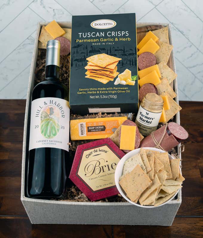 a gift basket with contents laid out across a white wood table. A bottle of red wine, a glass of red wine, a block of cheddar cheese, a summer sausage, a jar of honey mustard, a box of crackers, with a cutting board of sliced meat and cheese 