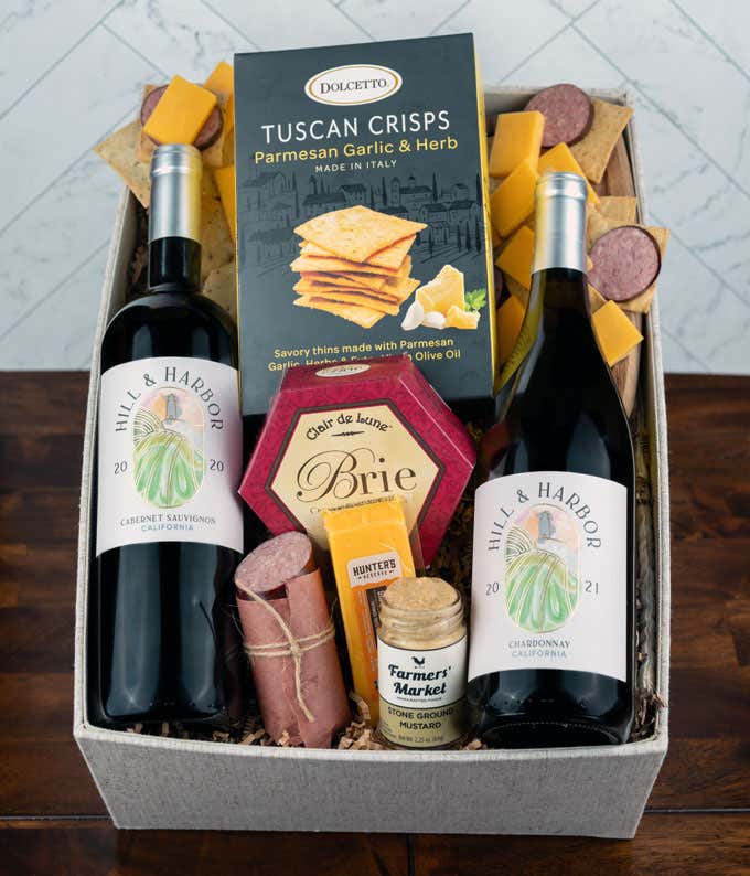 a gift basket with contents laid out across a white wood table. A bottle of red wine, a glass of red wine, a bottle of white wine, a glass of white wine, a block of cheddar cheese, a summer sausage, a jar of honey mustard, a box of crackers, with a cuttin