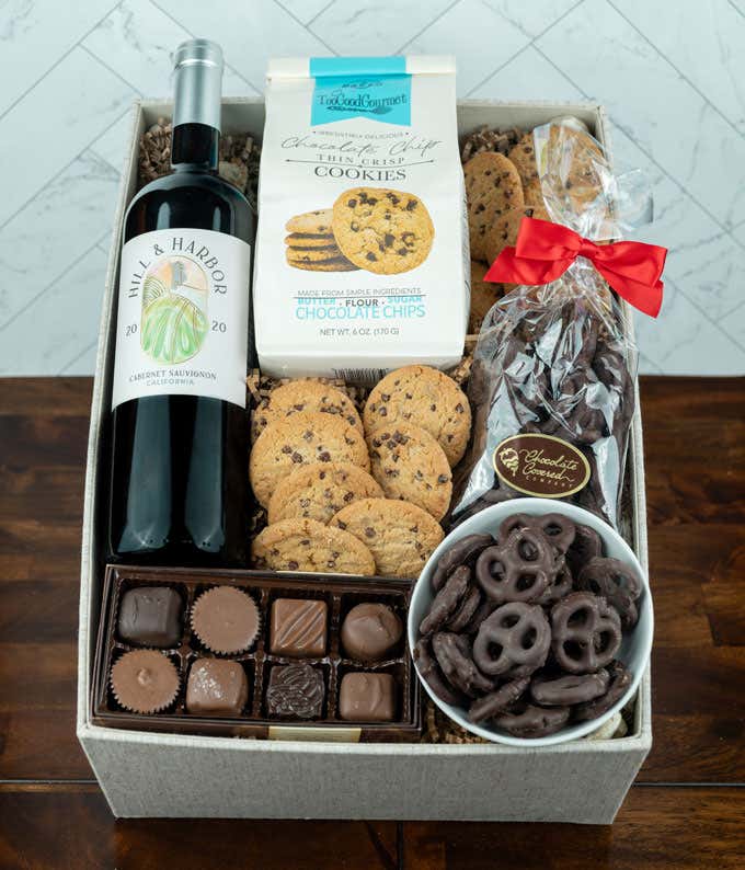 a bottle of red wine, two glasses of the wine, a bag of milk chocolate pretzel balls, a box of chocolate chip cookies, a box of assorted chocolates, and a cutting board all alid out on a white wooden table