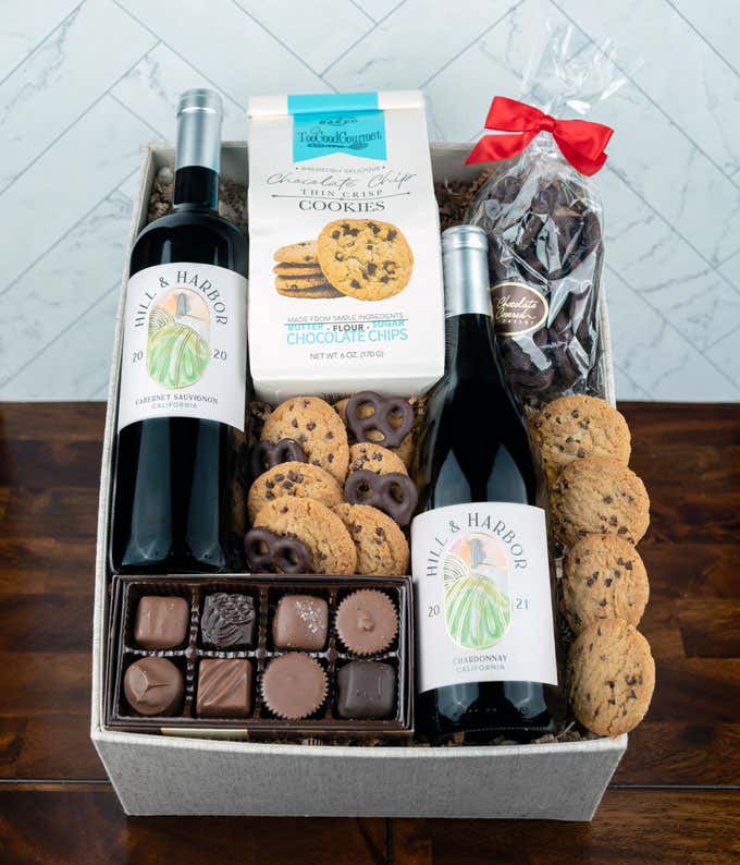 a bottle of white wine, a bottle of red wine, two glasses of the wine, a bag of milk chocolate pretzel balls, a box of chocolate chip cookies, a box of assorted chocolates, and a cutting board all alid out on a white wooden table