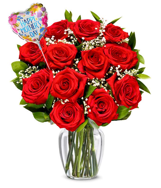 One dozen red roses with pops of greenery and white baby's breath in a clear glass vase with a pink and red heart shaped balloon reading happy mother's day