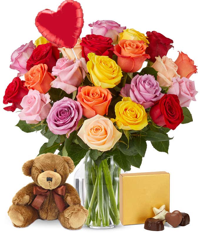 Two dozen roses red, orange, yellow, cream, pink, and lavender blooms with green leaves in a clear vase. Sat next to it is a brown teddy bear and a gold square box of chocolates with a red heart shaped balloon
