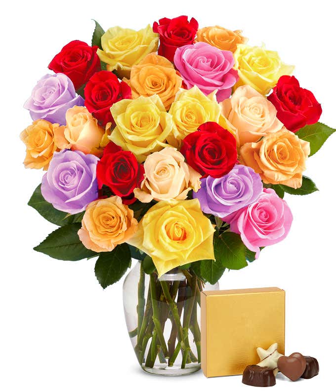 Mixed bouquet of vibrant roses in red, yellow, pink, orange, lavender, and cream displayed in a clear glass vase, accompanied by a gold box of assorted chocolates.