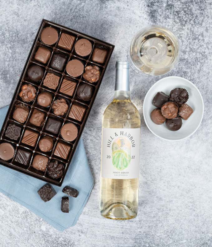 a bottle of white wine and a box of assorted chocolates laid out on a table with glasses of wine, cloth napkins, loose chocolates, and a cutting board