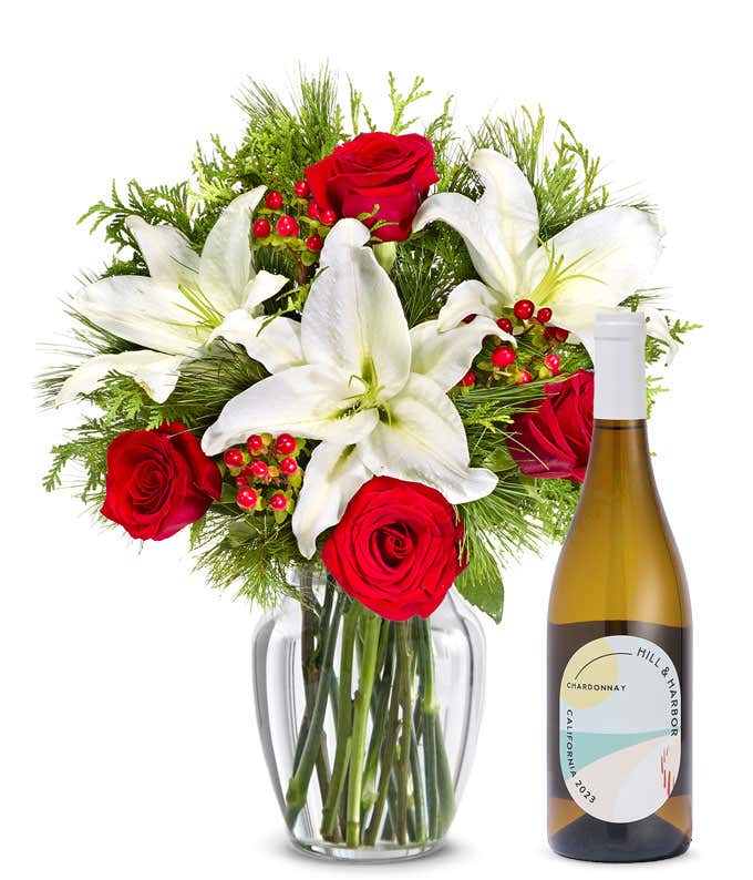  Bouquet of red roses, white lilies, greenery, and red berries arranged in a clear glass vase, accompanied by a bottle of Hill & Harbor Chardonnay.