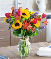 Alternative image 1 for A vibrant arrangement featuring sunflowers, pink tulips, red alstroemeria, purple Monte Casino asters, and green poms, optionally arranged in a glass vase. This lively mix offers a bright and cheerful display.