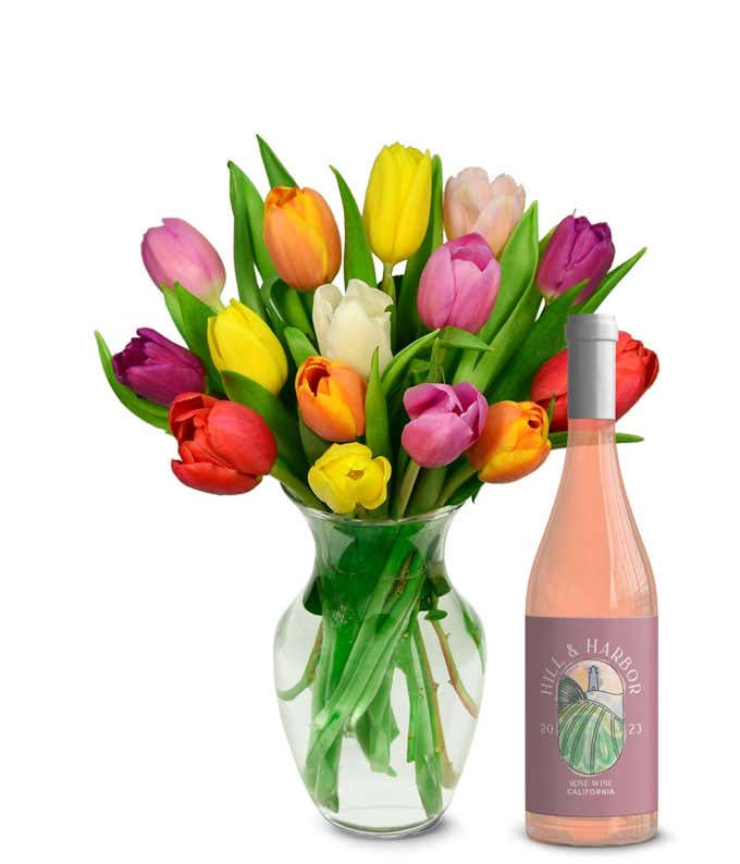 A vibrant array of tulips in red, pink, yellow, orange, and white, each bloom vivid and fresh, stands in a clear glass vase. Accompanying the flowers is a bottle of rose wine with a minimalist pink label, indicating a 2023 vintage from California.