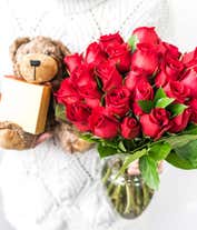 Alternative image 3 for a bouquet of 24 red roses with small white flowers and greenery in between in a clear glass vase with a small gold box of chocolates and a small brown teddy bear next to it