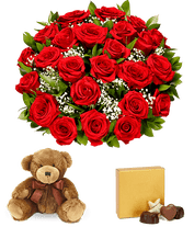a bouquet of 24 red roses with small white flowers and greenery in between in a clear glass vase with a small gold box of chocolates and a small brown teddy bear next to it