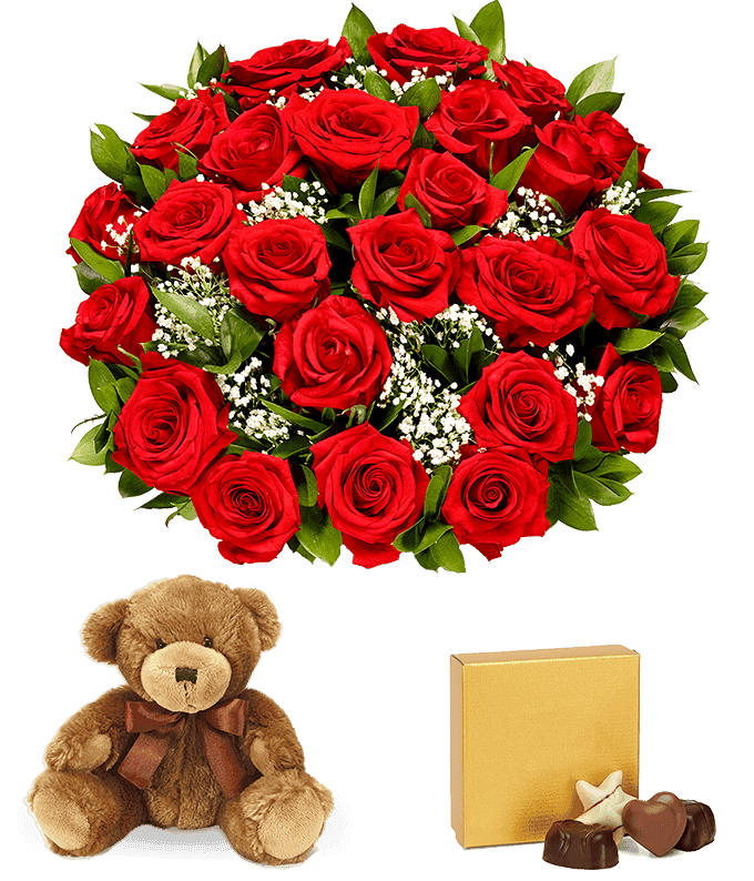 a bouquet of 24 red roses with small white flowers and greenery in between in a clear glass vase with a small gold box of chocolates and a small brown teddy bear next to it