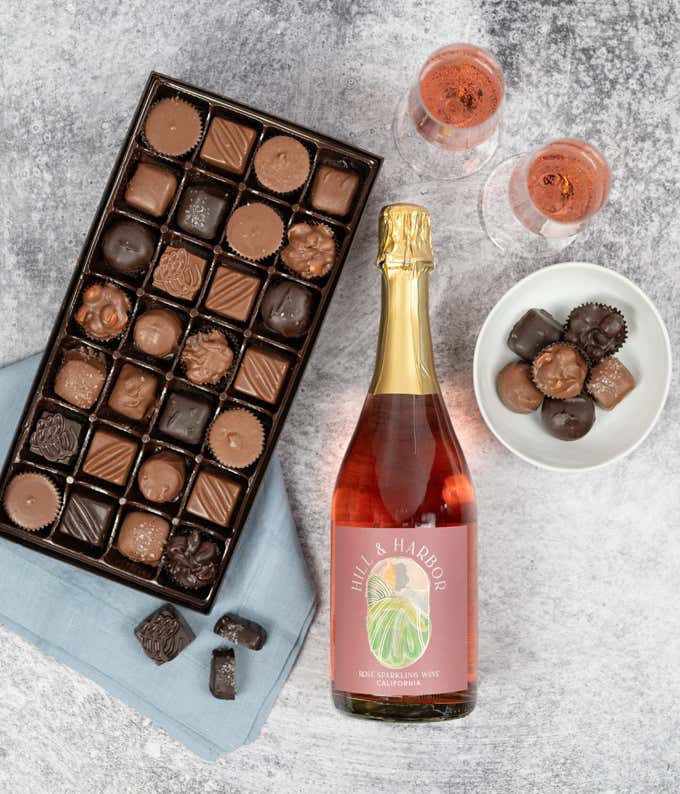 overhead view of an open box of 28 assorted milk and dark chocolates and a bottle of rose wine. There are also 2 glasses of rose, a small white ceramic bowl of chocolates, and a blue cloth napkin.