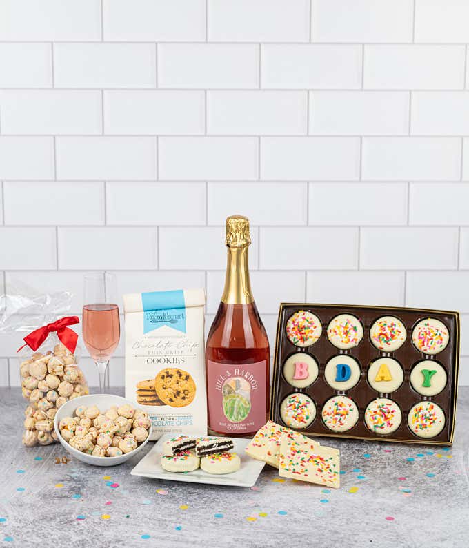 a bottle of pink sparkling wine, 12 white chocolate oreos, 4 topped with B-D-A-Y and 8 topped with rainbow sprinkles. A bag of chocolate chip cookies, a bag of whte chocolate and sprinkle covered popcorn, a glass of wine, white ceramic bowls and plates of
