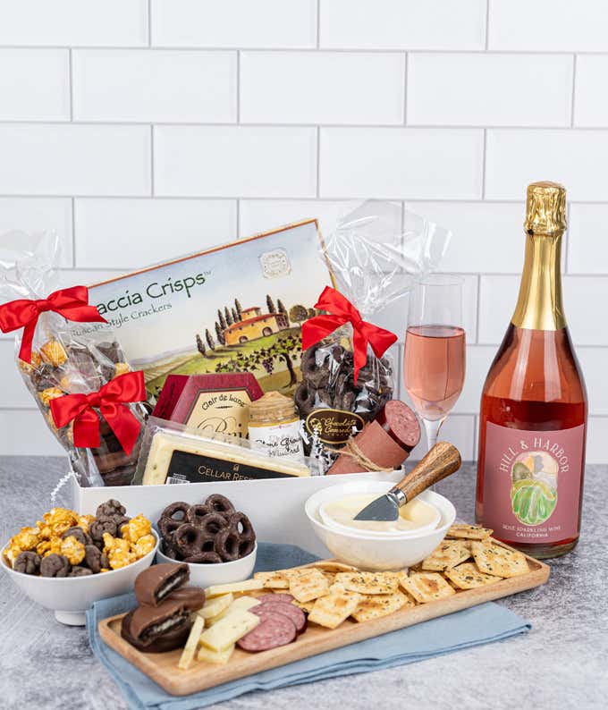 a gift box filled with crackers, cheeses, mustard, sausage, chocolate pocorn, and chocolate oreos on a kitchen counter with a bottle of sparkling pink wine