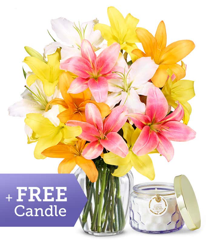 A clear glass vase filled with a vibrant bouquet of lilies in shades of pink, yellow, white, and orange, accompanied by a dahlia and orchid scented candle in a glass jar.