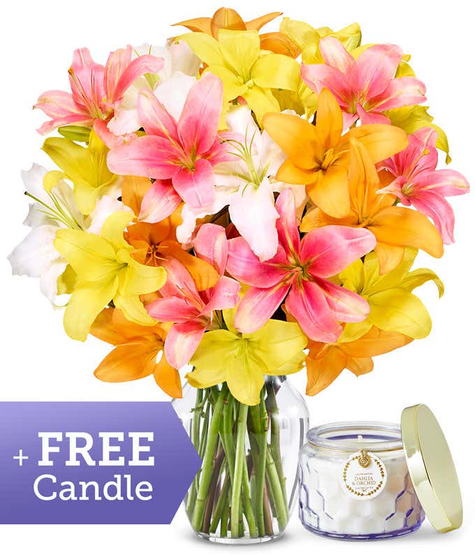 A clear glass vase filled with a large, colorful bouquet of lilies in pink, yellow, orange, and white, displayed alongside a dahlia and orchid scented candle in a clear glass jar.
