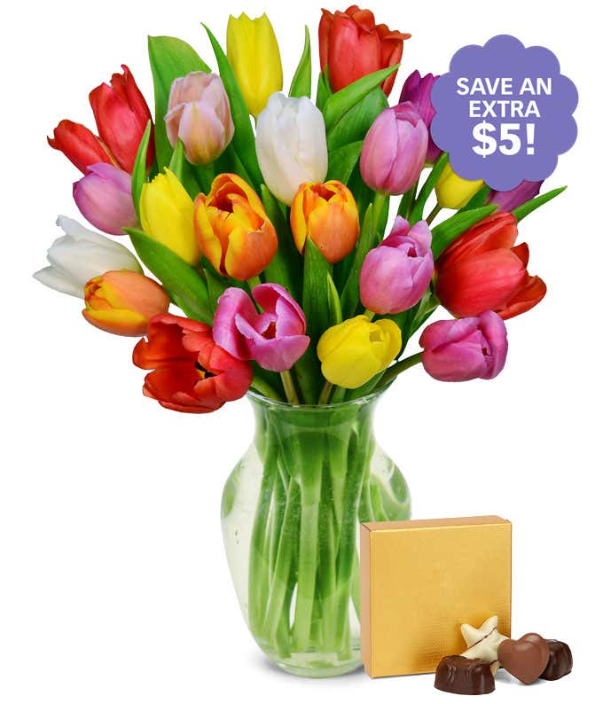 A clear glass vase filled with a colorful bouquet of multicolored tulips in red, yellow, orange, pink, purple, and white, displayed alongside a box of chocolates.