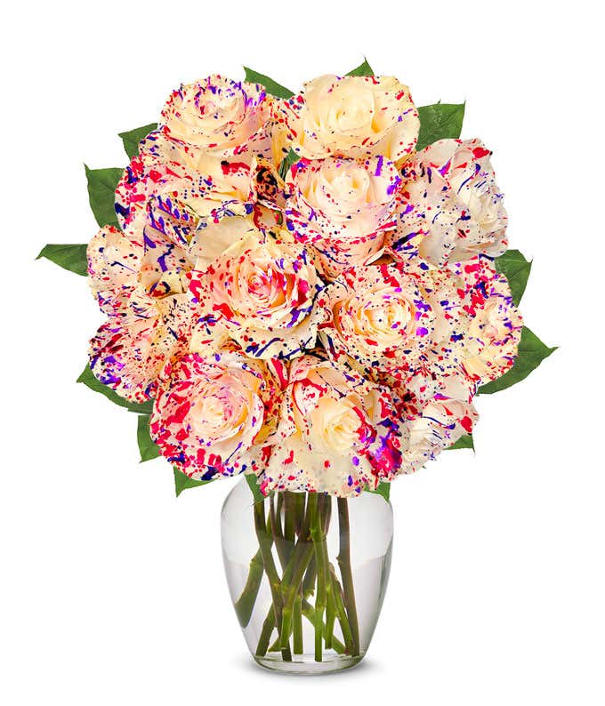A bouquet of cream roses splattered with bright red, purple, and pink paint, arranged in a clear glass vase with green leaves.