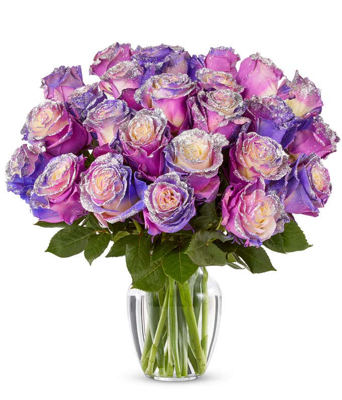 A bouquet of pink, purple, and cream roses with glittered petal edges arranged in a clear glass vase with green leaves.