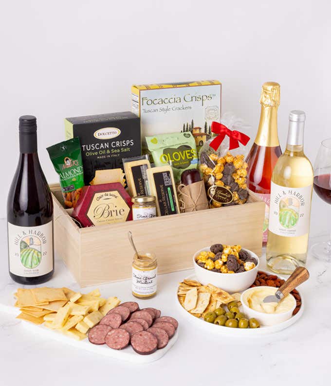 A gourmet gift basket with red, white, and sparkling ros wines, Tuscan crisps, assorted cheeses, crackers, olives, mustard, nuts, caramel popcorn, and salami, presented in a wooden tray with various dips and snacks.