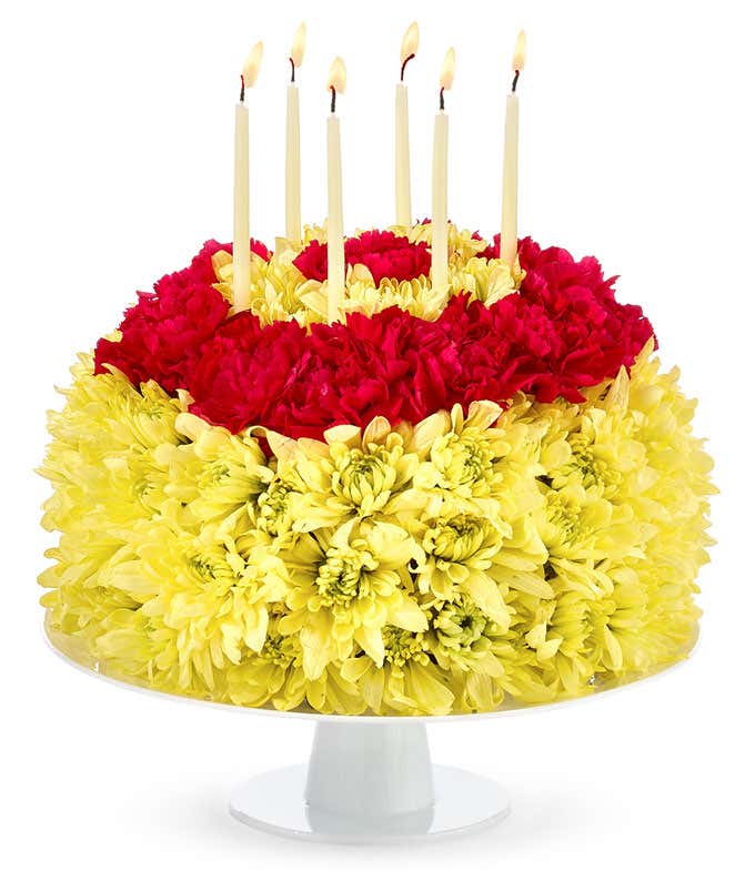 Floral arrangement made to look like a cake with red carnations and yellow poms and candles atop the cake