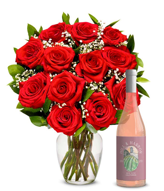 A bouquet of vibrant red roses arranged in a clear glass vase, surrounded by small white flowers and lush green leaves, paired with a bottle of rose wine from California, 2023, featuring a stylish, minimalist label.