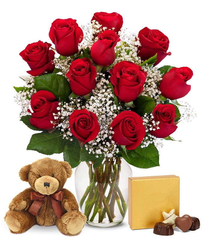 a bouquet of 12 red roses with small white flowers and greenery in between in a clear glass vase with a small gold box of chocolates and a small brown teddy bear next to it
