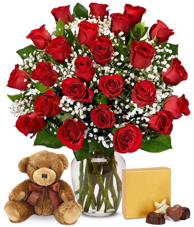 a bouquet of 24 red roses with small white flowers and greenery in between in a clear glass vase with a small gold box of chocolates and a small brown teddy bear next to it