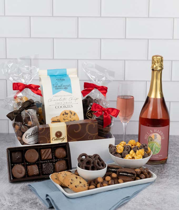 a gift box and a bottle of sparkling wine on a kitchen counter with a glass of wine and some chocolate treats on plates. Gift box includes cookies, chocolates, peanut butter chocolates, chocolate oreos, and chocolate pretzels