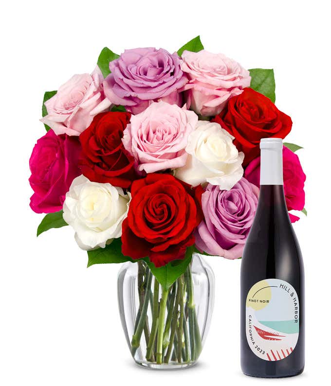 One dozen pink, purple and red roses arranged in a clear glass vase sits beside a bottle of Hill & Harbor California Pinot Noir.