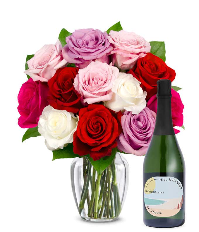 One dozen pink, purple and red roses arranged in a clear glass vase sits beside a bottle of Hill & Harbor California Sparkling Wine.