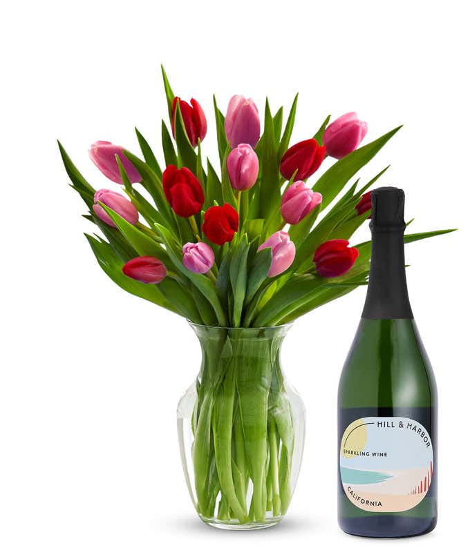A clear glass vase filled with 15 stems of mixed red and pink tulips and bright green stems sits beside a bottle of Hill & Harbor California Sparkling Wine.