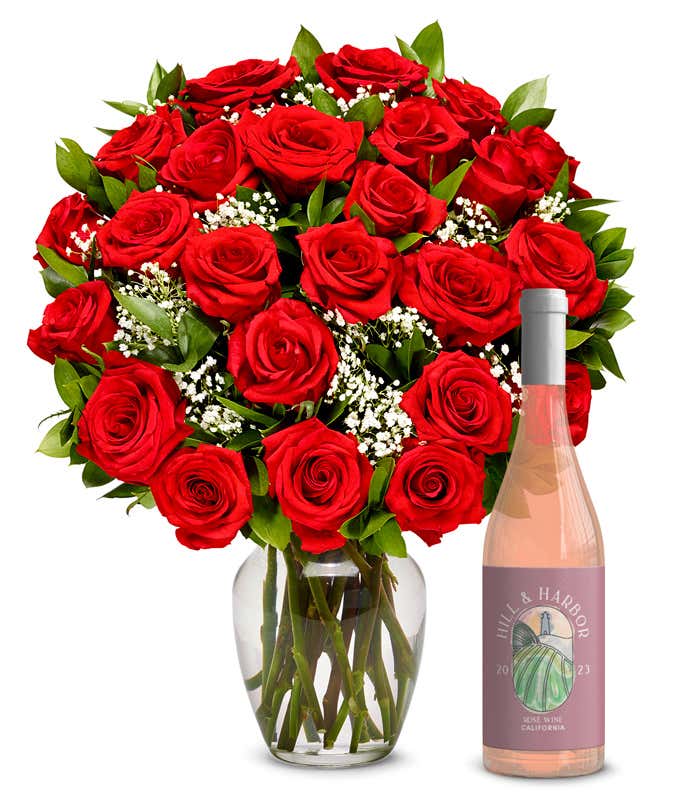 A lush bouquet of red roses, with deep velvet petals, nestled in a clear vase among tiny white blossoms and green leaves, complements a sleek rose wine bottle, vintage 2023 from California, radiating elegance and romance.