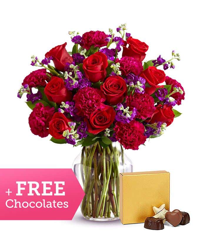 A bouquet of red roses, red carnations, and purple stock in a glass vase with a box of chocolates.