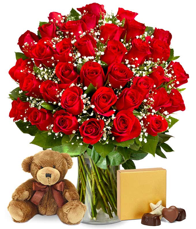 Image of 36 red roses beautifully arranged in a glass cylinder vase, accompanied by a box of chocolates and a cuddly bear plush, the ideal gift for conveying love and warmth.