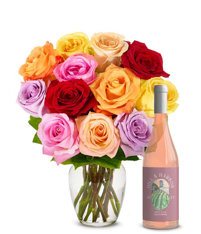 A bouquet of roses in various colors including red, pink, orange, yellow, and purple, neatly arranged in a clear glass vase. Beside it is a slender bottle of rose wine with a pale pink label from California, 2023, showcasing an elegant design.