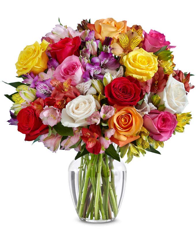 A bouquet of multicolored roses and alstroemeria in shades of red, pink, yellow, orange, purple, and white arranged in a clear glass vase.