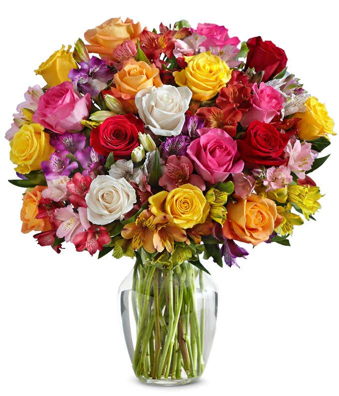 A bouquet of multicolored roses and alstroemeria in shades of red, pink, yellow, orange, purple, and white arranged in a clear glass vase.