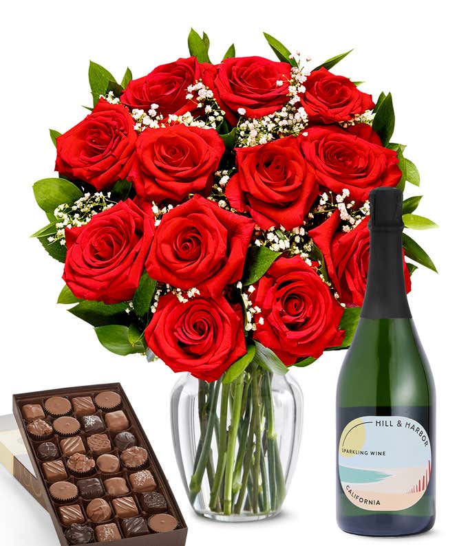 A lush arrangement of a dozen red roses in a clear vase sits beside a bottle of Hill & Harbor Sparkling Wine and a 1-pound box of gourmet assorted chocolates.
