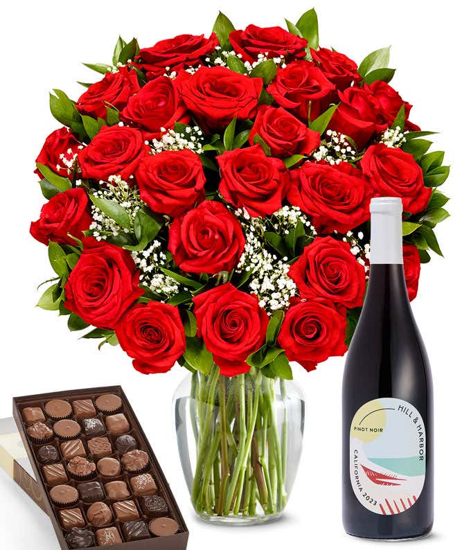 A lush arrangement of 2 dozen red roses in a clear vase sits beside a bottle of Hill & Harbor Pinot Noir and a 1-pound box of gourmet assorted chocolates.