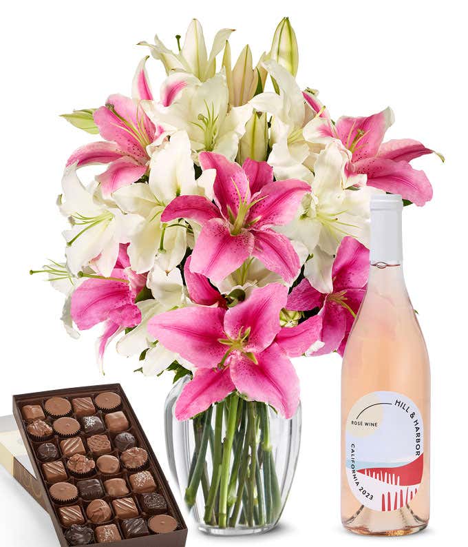 A bouquet of pink and white lilies arranged in a clear vase sits beside a bottle of Hill & Harbor Rosé and box of assorted chocolates.