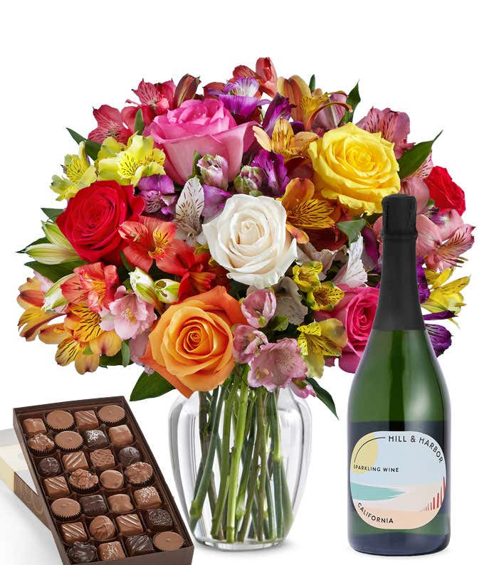 A colorful mixed bouquet of roses and alstroemeria flowers arranged in a clear vase is displayed beside a bottle of Hill & Harbor Sparkling Wine and a box of assorted chocolates.
