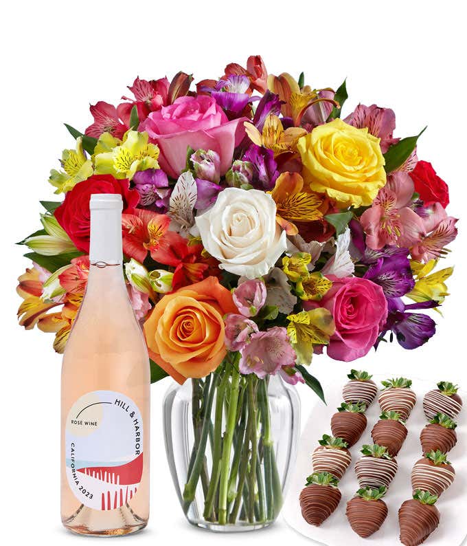 A bouquet of colorful roses and alstroemeria in a clear vase is displayed beside a bottle of Hill & Harbor Rosé and 12 chocolate-covered strawberries.