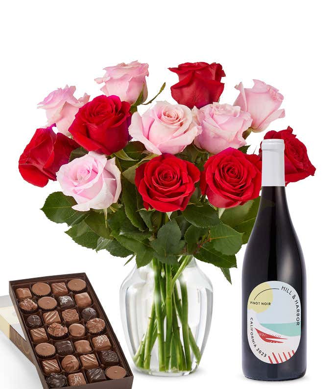 A bouquet of 12 red and pink roses arranged in a glass vase is displayed beside a bottle of Hill & Harbor Pinot Noir and a 1 pound box of assorted chocolates.