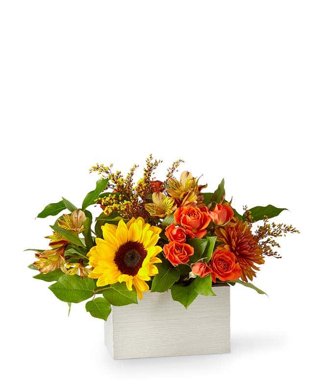 A vibrant floral arrangement featuring orange spray roses, yellow alstroemeria, butterscotch mums, red-tinted solidago, sunflowers, deluxe and premium orange lilies, and seasonal greens, all beautifully arranged in a white wooden box.