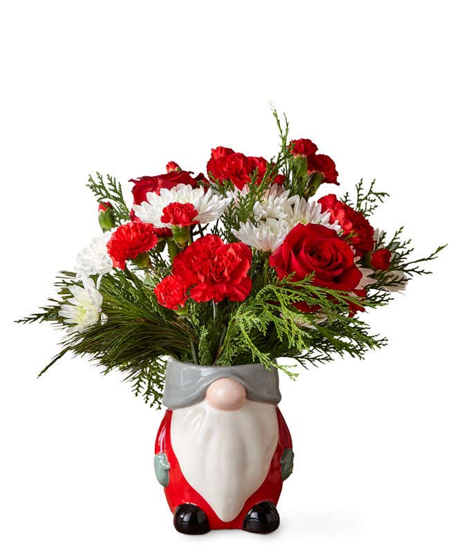 Red and white flowers arranged with assorted Christmas greens, placed into a festive gnome shaped ceramic vase
