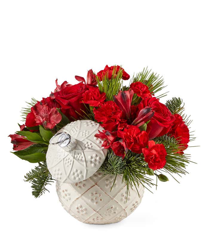 Red roses, red carnations, and red alstroemeria arranged with mixed Christmas greens, in a white ceramic ornament container