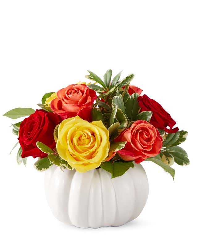 A white pumpkin filled with red, yellow, and orange roses, with greens throughout