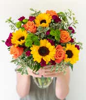 Alternative image 1 for A floral arrangement featuring orange roses, sunflowers, violet mini carnations, and green button poms in a clear vase.