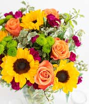 Alternative image 3 for A floral arrangement featuring orange roses, sunflowers, violet mini carnations, and green button poms in a clear vase.
